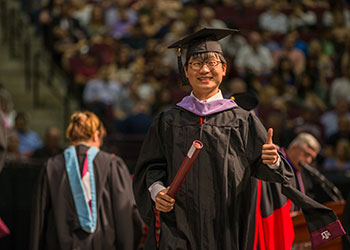 Texas A&M University student wearing graduation regalia, holding a diploma and giving a thumbs up sign with his hands.