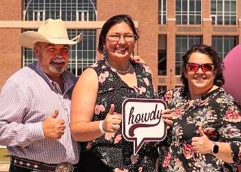 Three Aggie parents standing in front of Kyle Field, giving the thumbs up sign with their hands and holding a sign that says Howdy.